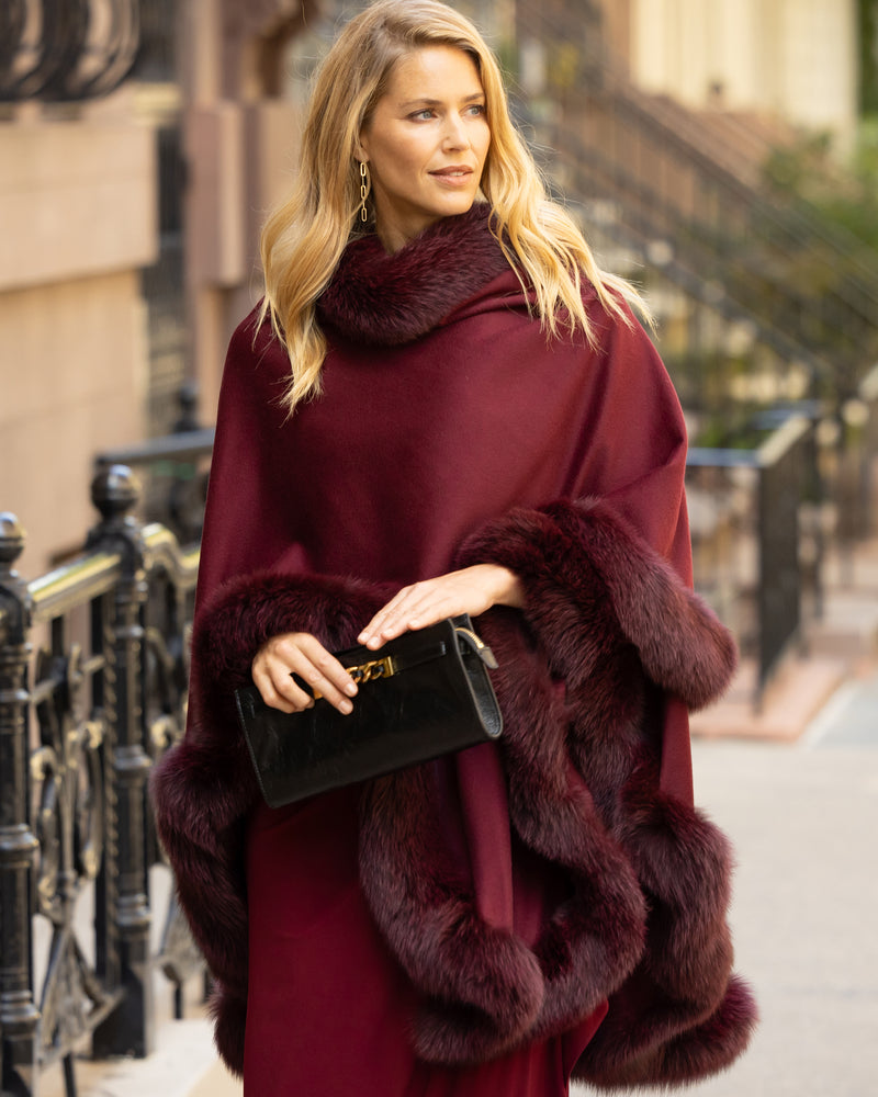 Woman wearing a fur trimmed cashmere cape coat in burgundy and holding a clutch handbag on a city street.