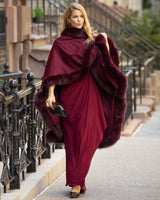 Woman in a burgundy dress fur trimmed cashmere cape in burgundy walking on a city street.
