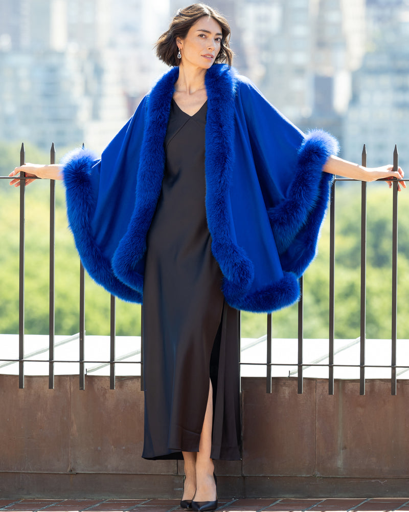 Woman wearing a Cervinia Cashmere Cape with real fur trim in Electric Blue over silk dress with a cityscape background