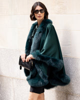 Woman wearing a Fur Trimmed Cape in Emerald against a stone wall.