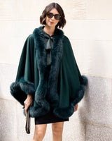 Woman wearing a Fur Trimmed Cape in Emerald against a wall.