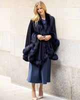 Woman wearing a a fur trimmed cape coat in navy against a light wall.