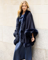 Woman wearing a cashmere cape with fur trim in navy against a light wall.