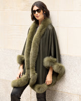 Woman wearing a Cashmere cape coat with real Fur trim in Olive against a stone wall.