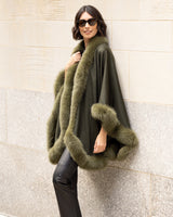 Woman wearing a Cashmere cape coat with real Fur trim in Olive against a stone wall.