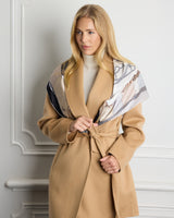 A blonde woman models a camel-colored wool coat with a matching tie belt, styled with a patterned beige and scarf draped over her shoulders, set against a white paneled wall.