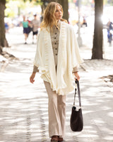 Woman walking outdoors on city sidewalk wearing a Knitted Cashmere Cape in Ivory with ruffles, plaid shirt, and plain pants with a handbag.