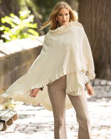 Woman wearing a Knitted Cashmere Cape in Ivory with ruffles wrapped over her outdoors on sidewalk with trees in the background
