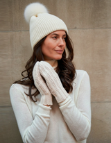 Woman wearing Montello Ribbed Cashmere Pompom Beanie in ivory with a pom-pom, gloves, and sweater against a beige wall.