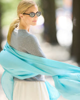 A woman wearing round sunglasses, and a  knit sweater with a flowing turquoise scarf wrapped around her shoulders, set against outdoor background.