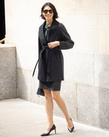 Woman wearing the Victoria cashmere belted coat in black and skirt set against a light stone wall.